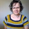 Smiling woman with short curly hair and glasses, wearing a short sleeved crocheted Sunflower Road Tee with a square neckline, in stripes of mustard yellow, navy and light grey.