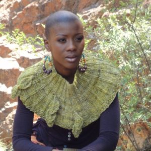 Leaves Cowl - A woman sitting in front of a rocky outcrop wearing a green knit cowl with knit leaves attached.