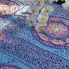 A crocheted blanket called At the Rainbow Bridge is laid out flat, featuring large circular floral motifs in pink, purple, yellow, and green. The squares are bordered with lace-like stitching in blue and purple. A white orchid with yellow centres rests gently on top of the blanket, adding a natural accent to the scene.