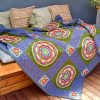 A wooden bench or daybed with beige cushions is topped with a colorful crocheted blanket called At the Rainbow Bridge. The blanket features large circular floral motifs in red, white, green, and blue, framed by green squares and bordered in purple. Smaller red and white squares accent the corners. The setting is warm and homey, with natural textures and soft lighting.