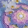 A close-up of crocheted mandala patterns in pastel shades of pink, purple, blue, yellow, and white, from the At the Rainbow Bridge Blanket. The designs feature layered, petal-like shapes radiating from central white floral motifs. Several white orchid flowers with yellow centres rest on top of the crochet work, adding a soft, natural detail.