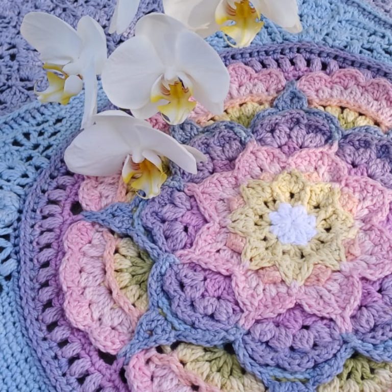 A close-up of crocheted mandala patterns in pastel shades of pink, purple, blue, yellow, and white, from the At the Rainbow Bridge Blanket. The designs feature layered, petal-like shapes radiating from central white floral motifs. Several white orchid flowers with yellow centres rest on top of the crochet work, adding a soft, natural detail.
