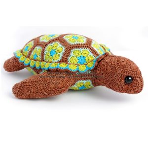 Snap the African Flower Turtle, designed by Heidi of Heidi Bears. Hand-crocheted stuffed turtle featuring a colorful granny square shell in earthy tones of brown, green, blue, and yellow. The shell is made of hexagonal and pentagonal motifs, and the head, legs, and tail are crocheted in solid brown yarn. Finished with black safety eyes, this soft toy showcases detailed craftsmanship and playful design.
