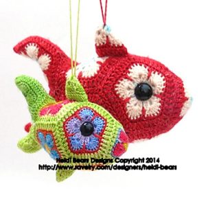 Two crocheted fish toys hang by strings. The larger fish is red with white and pink hexagons, and the smaller fish is green with multicoloured hexagons. Both have black button eyes and are crafted with textured crochet stitches.