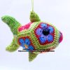A crocheted fish toy hangs by string. The fish is green with multicoloured hexagons. It has black button eyes and is crafted with textured crochet stitches.