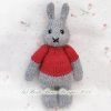Knitted grey bunny toy wearing a red sweater, lying on white floral fabric; handmade with stitched eyes and mouth.
