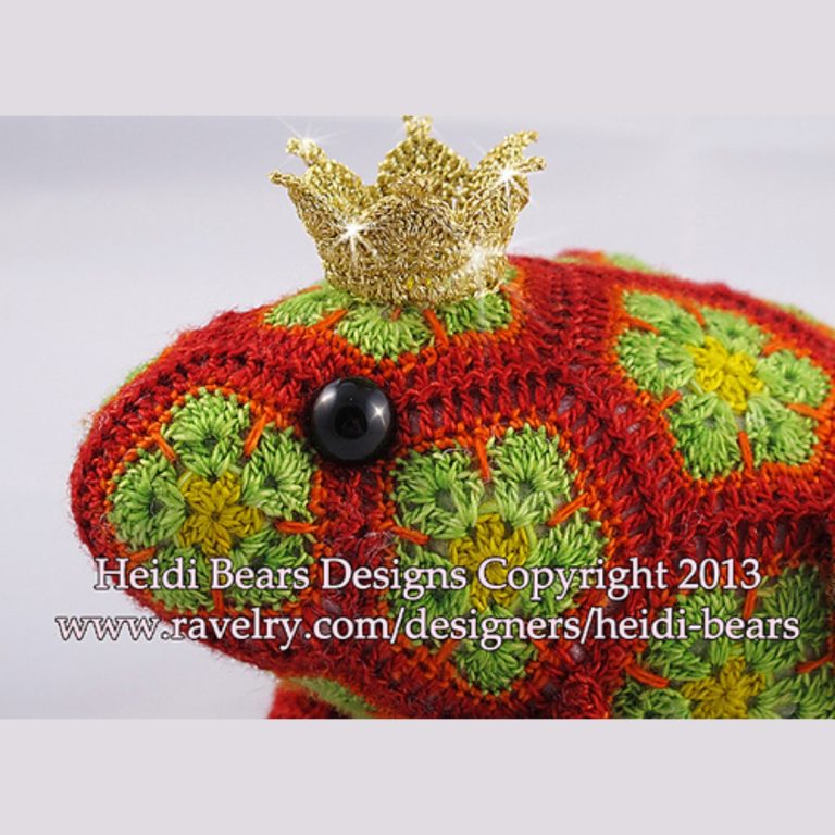 Close up of a crocheted frog prince toy made with red and green African flower motifs, featuring a golden crown and black button eyes; handcrafted using hexagons and pentagons pattern by Heidi Bears Designs.