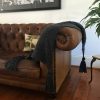 Chunky dark gray knitted throw with oversized tassels draped over a brown tufted leather sofa, styled with a nature-print cushion and leafy green plant in a warm, wood-floored interior.