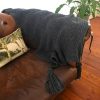 Textured dark gray knitted throw with oversized tassels draped over a brown leather couch, styled with a tropical palm tree cushion and leafy green plant on a wood floor for a warm, inviting look.