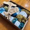 ColourSpun Tour de Fleece Mystery Box showing hand dyed Pure Merino, Kid Mohair and Tussah Silk fibre in teal and golden green tones, with a flat pack Niddy Noddy and Twist Gauge tool, packed in a kraft box.