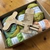 ColourSpun Tour de Fleece Mystery Box showing hand dyed Pure Merino, Kid Mohair and Tussah Silk fibre in soft green, cream and peach tones, with a flat pack Niddy Noddy and WPI measurement tool, packed in a kraft box.