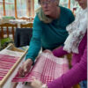 Two women working together at a rigid heddle loom, one holding scissors near red and white woven cloth while the other holds up the finished section of a handwoven tea towel.