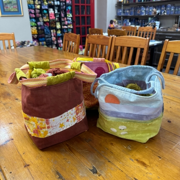 Three handmade project bags in rust, pink and pale blue with sunset appliqué, on a wooden table in the ColourSpun studio.