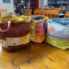 Three handmade project bags in rust, pink and pale blue, side by side on a wooden table showing their drawstring tops and handles.