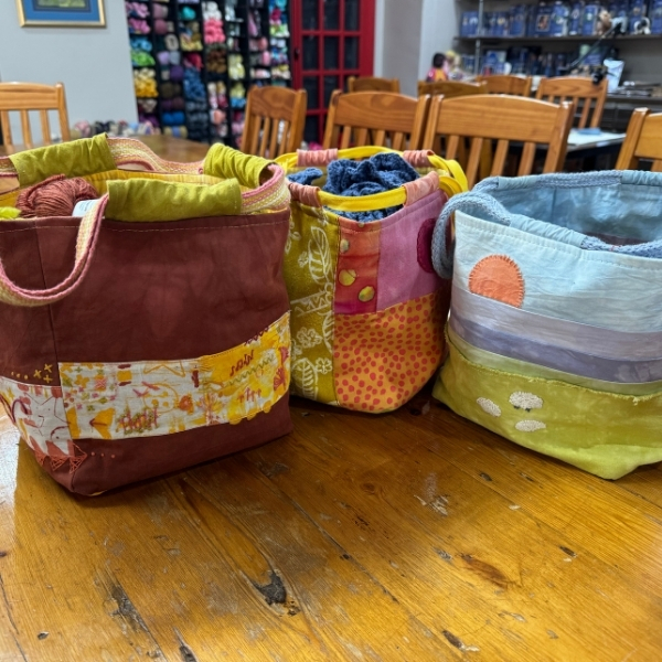 Three handmade project bags in rust, pink and pale blue, side by side on a wooden table showing their drawstring tops and handles.
