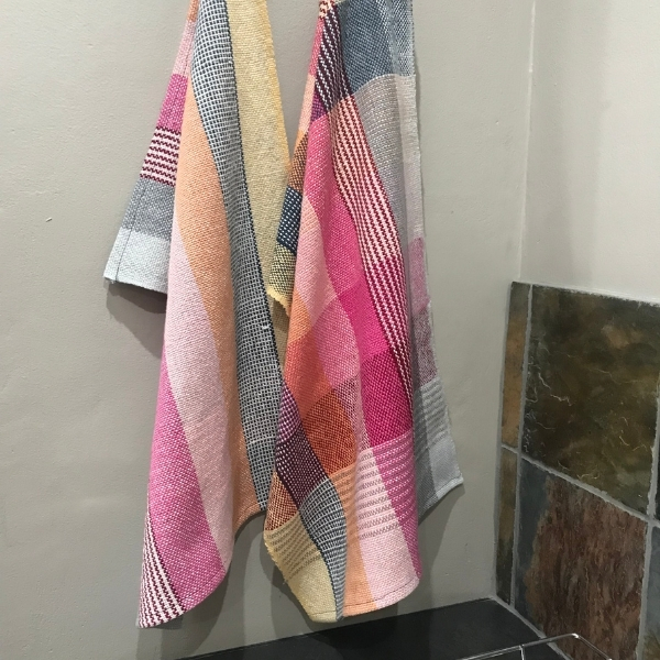 Two finished handwoven tea towels hanging on a hook, woven in a check pattern of pink, orange, yellow, grey and white.