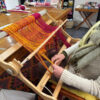 Close up of a rigid heddle loom warped with magenta and golden yellow yarn, hands tying on the ends ready to begin weaving, yarn on shelves in the background.