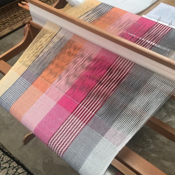 Overhead view of weaving in progress on a rigid heddle loom, showing a check pattern in pink, orange, yellow and grey, with a woven rug visible on the floor beneath the loom.