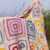 The finished Namaqua Blanket in cream with colourful granny squares, draped over dune grass with the sky behind
