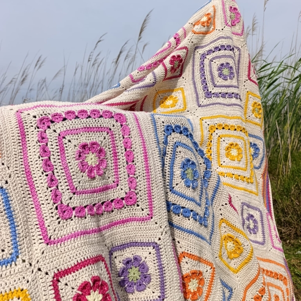 The finished Namaqua Blanket in cream with colourful granny squares, draped over dune grass with the sky behind