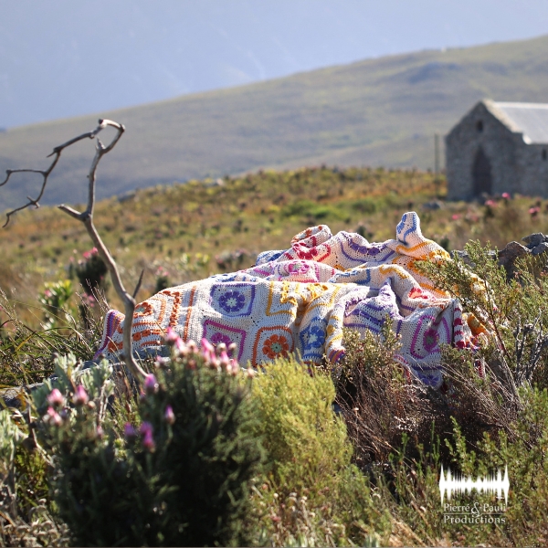 The Namaqua Blanket in cream with red, pink, blue, orange and purple diamond patterns, bundled among fynbos shrubs and pink erica flowers, with a bare branch in the foreground and an old stone church on a hazy mountainside behind