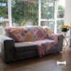 The Namaqua Blanket draped over a grey sofa in a sunlit living room, with green foliage visible through the window and a vase of yellow flowers on a side table