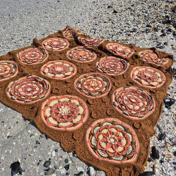 The Over the Coral Sea crochet blanket by Hooked on Sunshine in ColourSpun Pure Cotton DK, laid flat on a shell covered beach. Rows of round coral and cream mandala motifs are joined by caramel brown lacework and small raised roses.