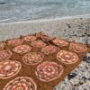 The Over the Coral Sea crochet blanket by Hooked on Sunshine, made in hand dyed ColourSpun Pure Cotton DK, spread out on a pebbly shell beach in bright sunshine with gentle waves behind it.