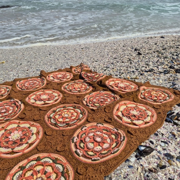 The Over the Coral Sea crochet blanket by Hooked on Sunshine, made in hand dyed ColourSpun Pure Cotton DK, spread out on a pebbly shell beach in bright sunshine with gentle waves behind it.