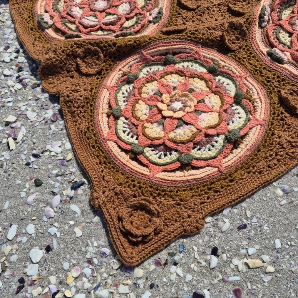 Close up of the Over the Coral Sea crochet blanket by Hooked on Sunshine, made in ColourSpun Pure Cotton DK. A round mandala motif in coral, blush pink, butter yellow, cream and sage green sits in a textured caramel brown border with a raised crochet rose, on sand scattered with small shells.