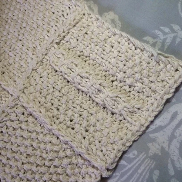 Cream cotton sample square from the Aran Caresse Cable Afghan Collection with a cable panel and a textured stitch, resting on grey patterned fabric