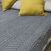 Grey Cable Me Cosy knitted blanket on a bed with chevron and lattice cable strips beside two mustard yellow cushions