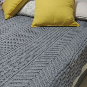 Grey Cable Me Cosy knitted blanket on a bed with chevron and lattice cable strips beside two mustard yellow cushions