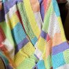 A crocheted Log Cabin Patchwork blanket by Hilda Steyn draped over a chair, made of rectangular blocks in lemon, mint, turquoise, lavender, coral, peach and cream, with some blocks in solid stitch and others in open lacy mesh.