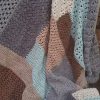 A crocheted Log Cabin Patchwork blanket by Hilda Steyn in muted tones, with blocks of grey purple, dusty pink, chocolate brown, pale aqua and cream, mixing dense stitches with open lacy ones.