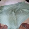Sage green Cable Me Cosy knitted blanket draped over a bed, showing cable strips, chevrons and a Celtic knot panel