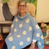 A smiling woman with short grey hair and glasses sits wearing the Daisies in Denim poncho by Hilda Steyn. It’s crocheted in soft denim blue with a cowl neck and joined square motifs, each with a white and yellow daisy at its centre.