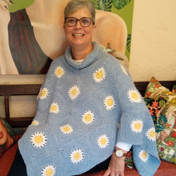 A smiling woman with short grey hair and glasses sits wearing the Daisies in Denim poncho by Hilda Steyn. It’s crocheted in soft denim blue with a cowl neck and joined square motifs, each with a white and yellow daisy at its centre.