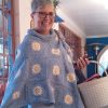 A smiling woman with short grey hair and glasses stands on a covered veranda wearing the Daisies in Denim poncho by Hilda Steyn. The denim blue crochet poncho has a cowl neck and white and yellow daisy motifs scattered across it.