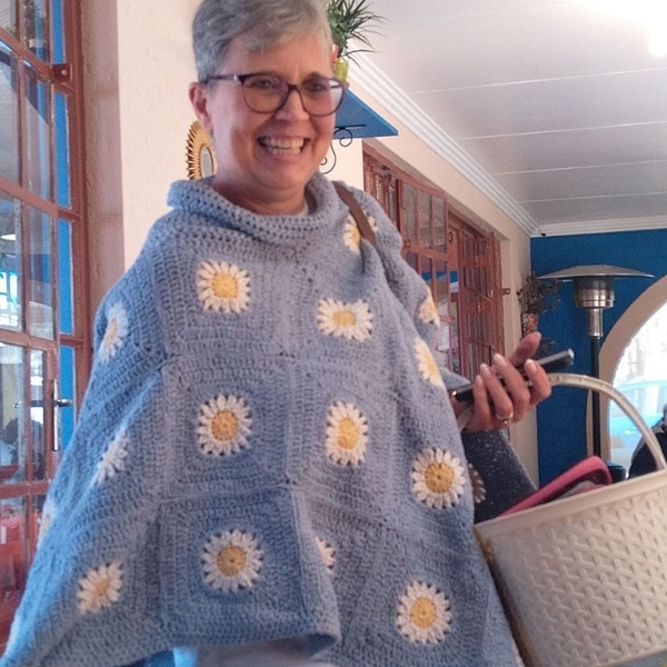 A smiling woman with short grey hair and glasses stands on a covered veranda wearing the Daisies in Denim poncho by Hilda Steyn. The denim blue crochet poncho has a cowl neck and white and yellow daisy motifs scattered across it.