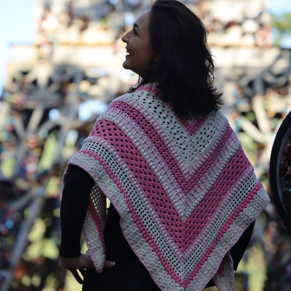 Smiling woman looking up and away, wearing the Ireland Shawl in pink and soft grey stripes against a blurred, colourful background.