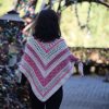 Back view of a woman wearing the Ireland Shawl, with pink and cream chevron stripes pointing down her back, as she walks past padlock covered railings.