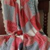 Patchwork blanket of textured squares in coral, blush pink, soft grey and cream, with a cream scalloped edge, draped over a dark wooden chair.