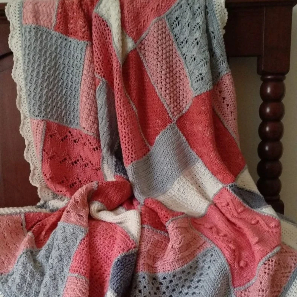 Patchwork blanket of textured squares in coral, blush pink, soft grey and cream, with a cream scalloped edge, draped over a dark wooden chair.