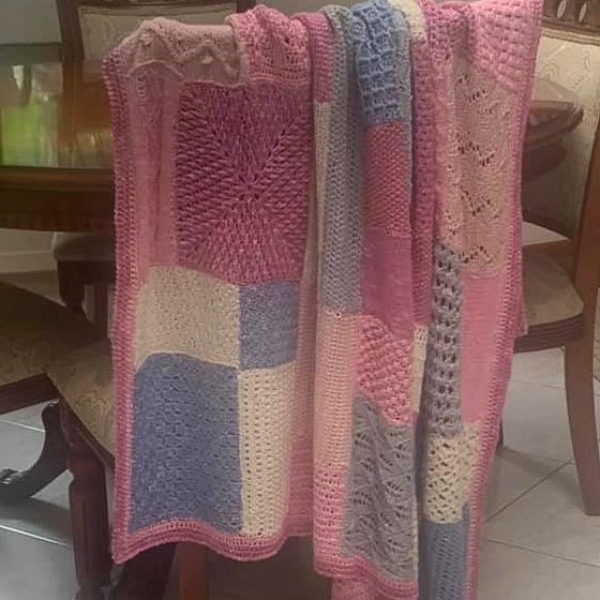 Patchwork blanket of stitch sampler squares in raspberry pink, rose, periwinkle blue and cream, with a pink border, draped over the back of a dining chair.