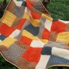 Patchwork blanket of textured squares in burnt orange, mustard yellow, caramel brown, taupe, slate blue and cream, with a yellow edging, spread out on green grass.