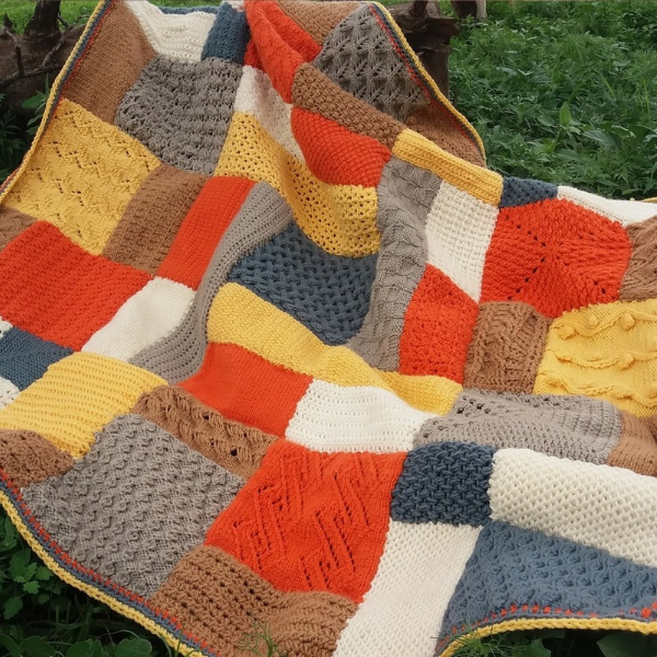 Patchwork blanket of textured squares in burnt orange, mustard yellow, caramel brown, taupe, slate blue and cream, with a yellow edging, spread out on green grass.
