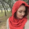 Woman looking down and to the side, wearing a bright red crocheted hooded cowl with a scalloped edge, standing among trees in soft misty light.