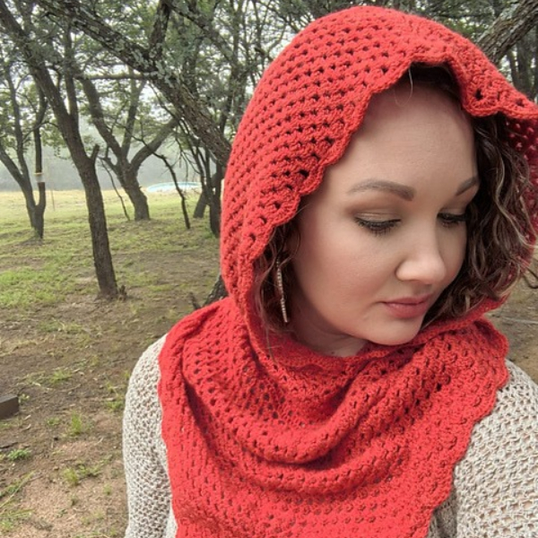 Woman looking down and to the side, wearing a bright red crocheted hooded cowl with a scalloped edge, standing among trees in soft misty light.