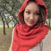 Woman with curly hair looking at the camera, wearing the red crocheted Rooikappie hood and cowl over a cream knit top, with trees and grass behind her.
